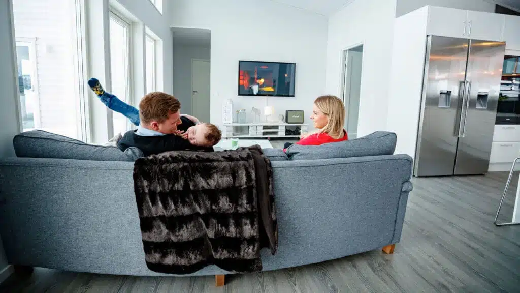 Två vuxna och ett barn sitter i en grå soffa sedda bakifrån och tittar på en väggmonterad TV; mörk pälspläd hänger över ryggstödet, Marshall-högtalare på en bänk och ett silverfärgat sidekylskåp till höger.