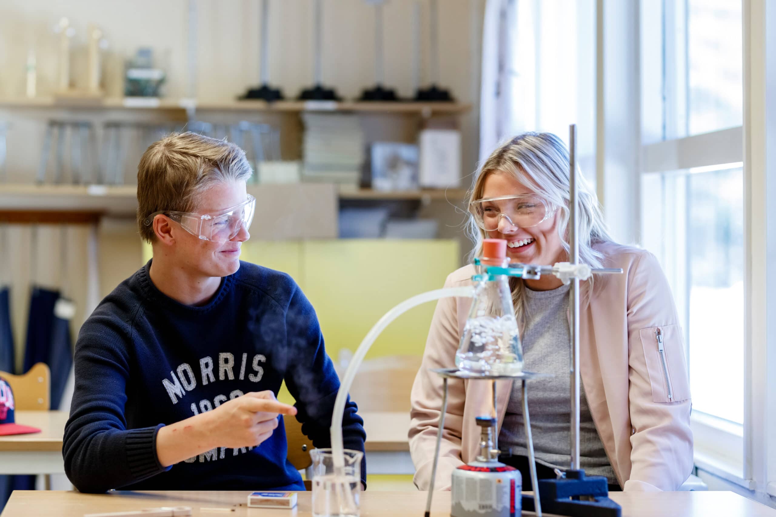 Två skrattande gymnasieelever med skyddsglasögon vid en labbänk; pojken i mörkblå Morris-tröja och flickan i ljusrosa jacka tittar på varandra bredvid en glaskolv på stativ.