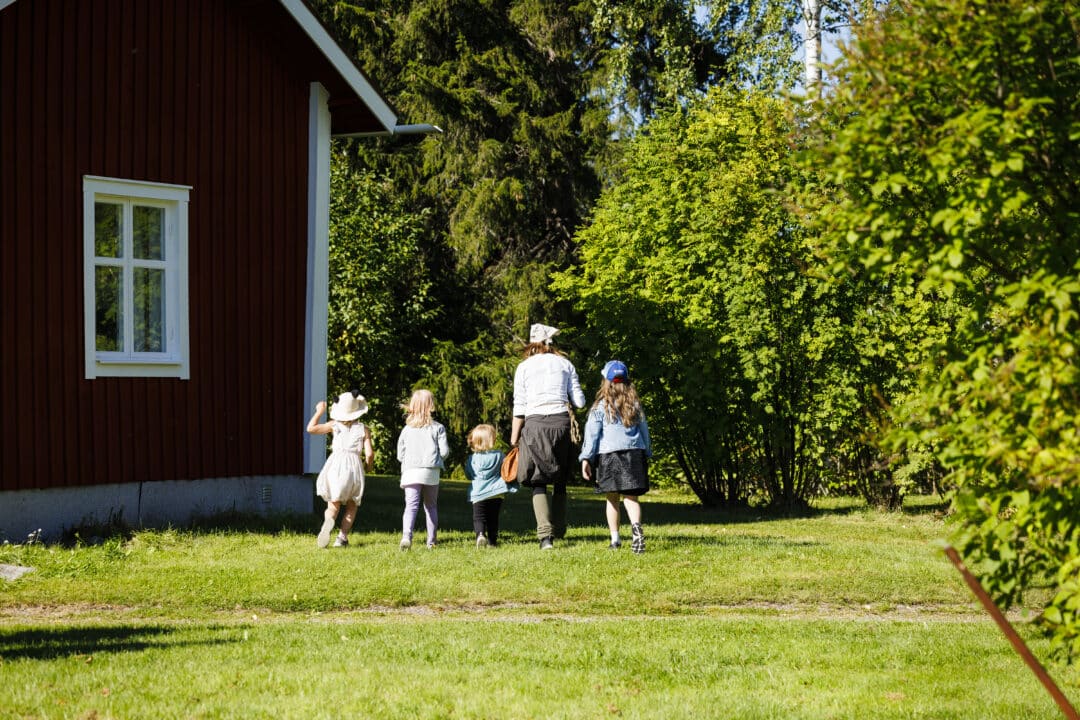 Four children and a woman is walking over a green lawn by a traditional red house.