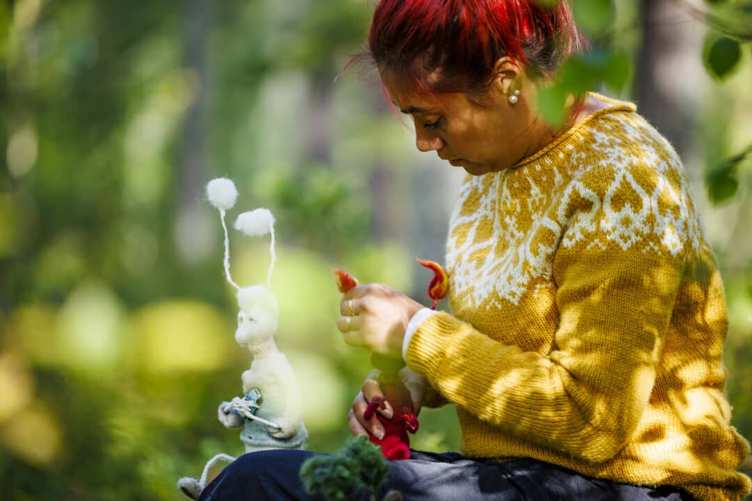 Woman in yellow knit shirt and red hais sits in forest and cragts wool figures. Ina runs her own creative company.