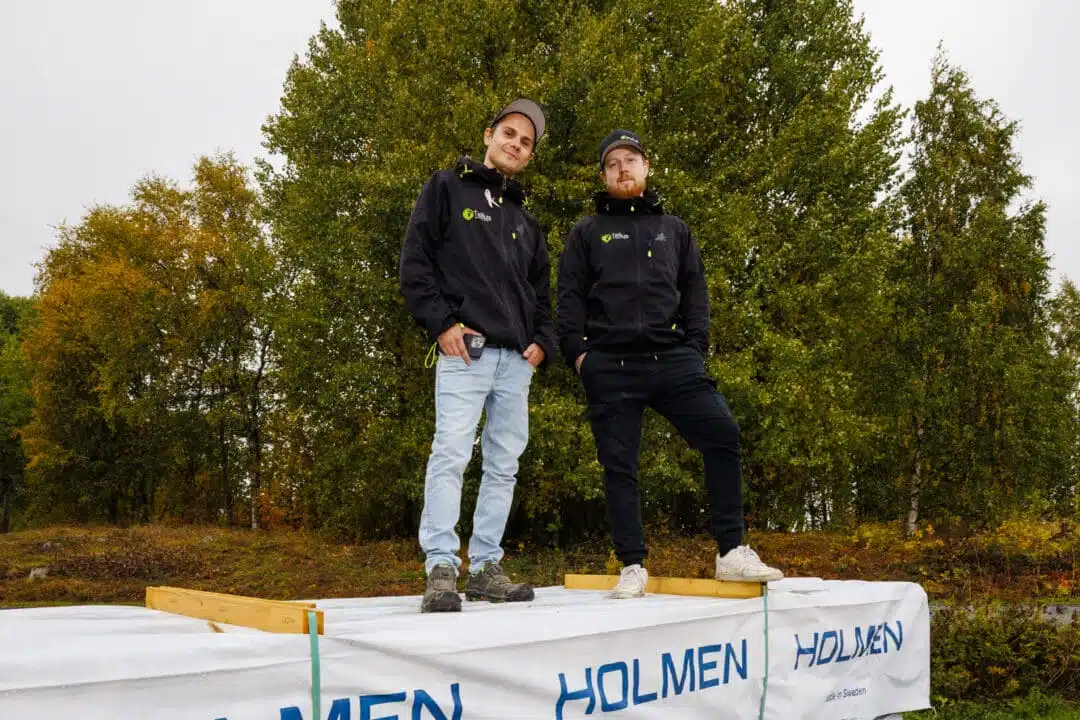 Två män i mörka Tellus-jackor och kepsar står ovanpå en stor virkesbunt märkt HOLMEN, omgivna av höstklädda björkar i grönt och gult.