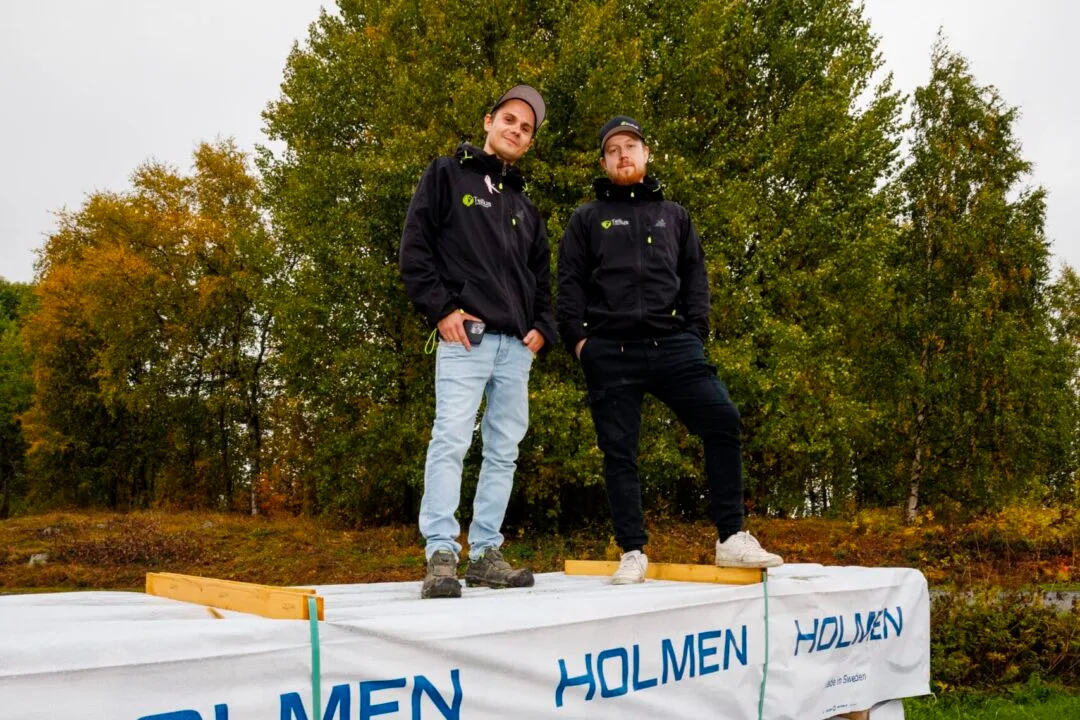 Två män i mörka Tellus-jackor och kepsar står ovanpå en trävirkesbunt märkt HOLMEN, omgivna av höstgula björkar under en grå himmel.