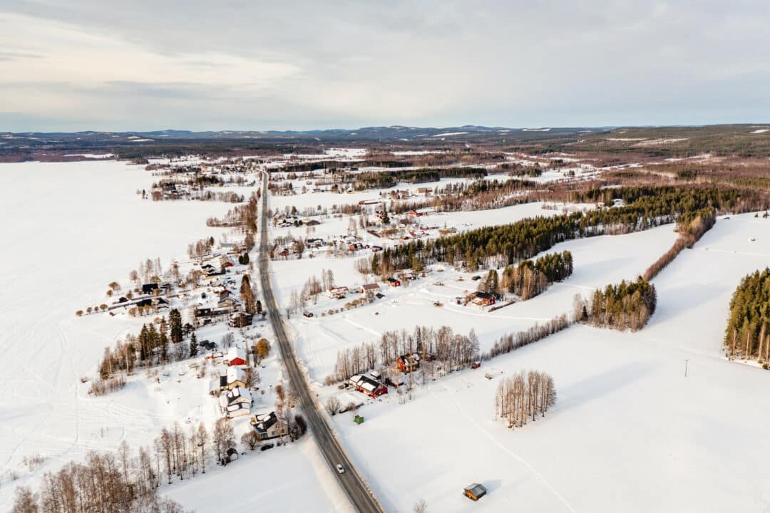Flygbild över ett snötäckt landskap där en rak landsväg löper genom utspridd bebyggelse omgiven av frusna fält, granskog och björkar i vinterskrud.