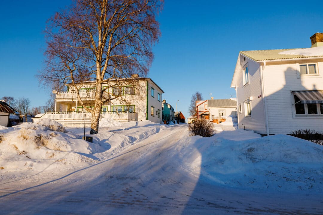 Snötäckt gata mellan en gulgrön tvåvåningsvilla med vit veranda och ett vitt trähus, under en klarblå himmel med kala lövträd.