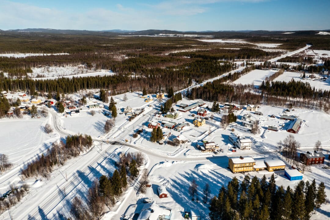 Flygbild över en vintrig by med röda, vita och gula hus tätt samlade längs vägar, snötäckta ängar och omgivande barrskog under blå himmel.