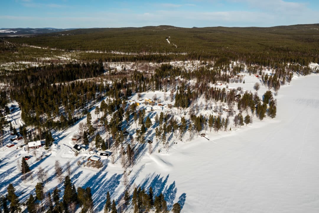 Flygbild över ett snötäckt landskap med spridda hus mellan barrskog vid en frusen sjö, omgivet av skogsklädda kullar under ljusblå himmel.