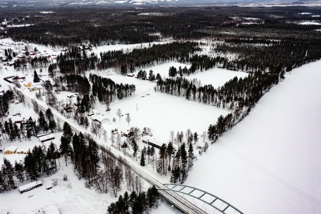 Flygbild en gråmulen vinterdag över en byggd bro över snötäckt älv, med skogsklädda områden, åkrar och spridda hus i bakgrunden.