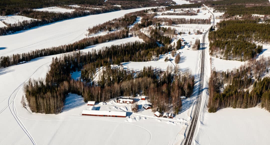Flygbild över en snötäckt by med röda gårdsbyggnader vid en väg, omgiven av vidsträckta vita fält, skogsdungar och spår efter skotrar.