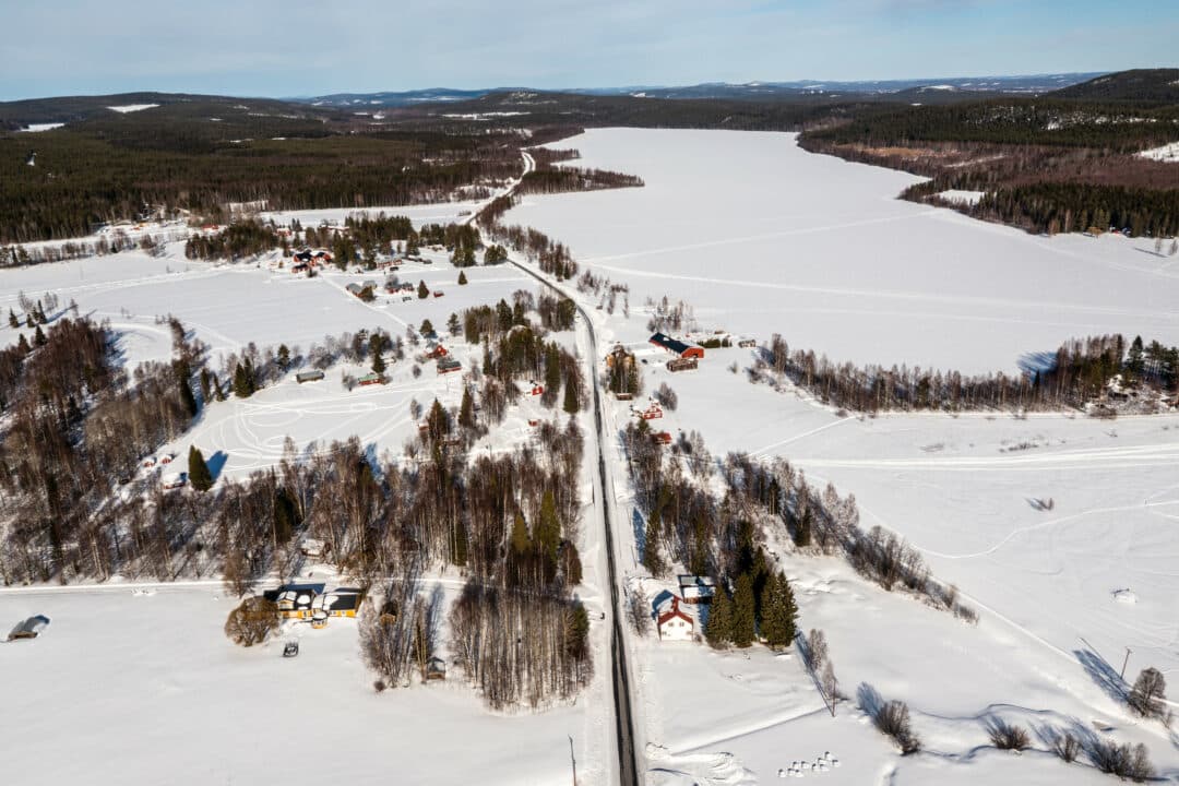 Flygbild över ett vinterlandskap med spridda gårdar längs en rak plogad väg, vidsträckta snötäckta fält och en frusen sjö omgiven av barrskog.