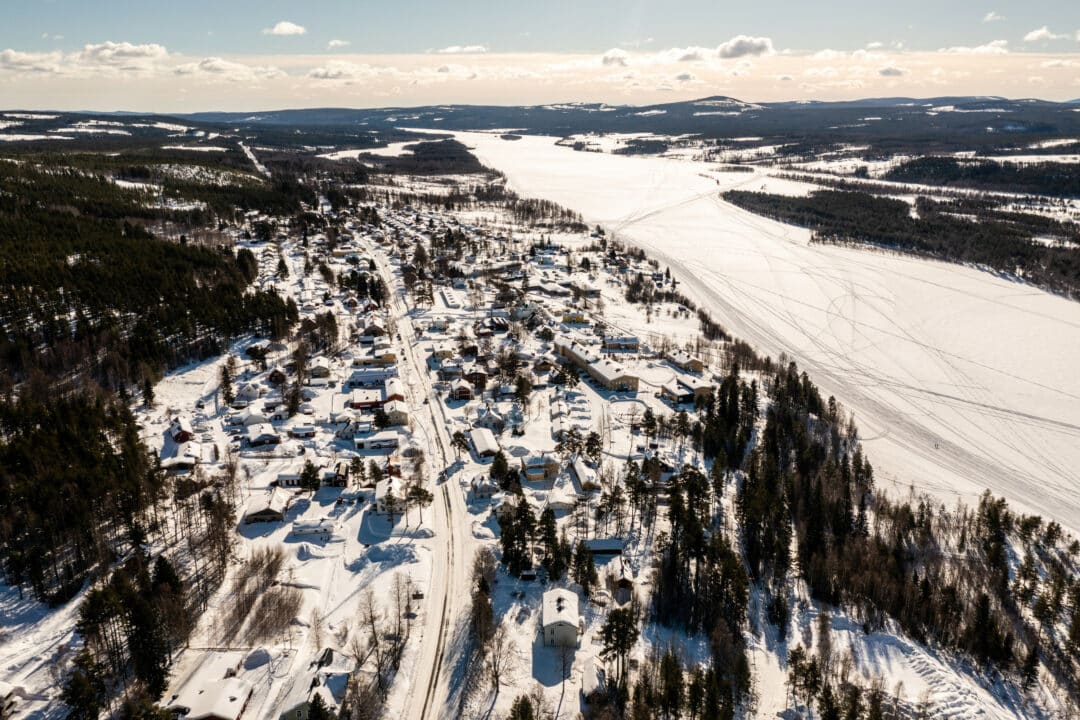 Flygbild över ett samhälle längs en bred frusen älv på vintern, med snötäckta hus, vägar och tät skog under en delvis molnig himmel.