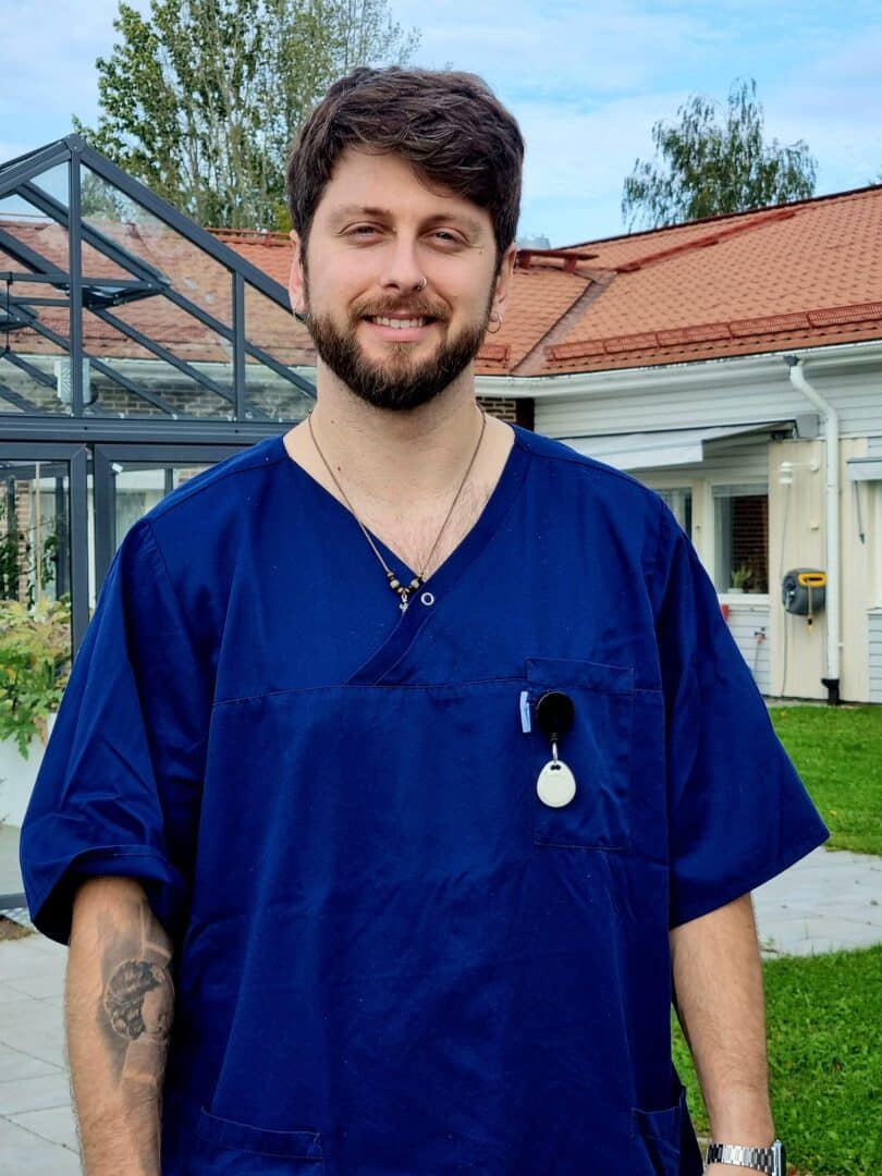 Leende skäggig man med mörkt hår klädd i mörkblå sjukvårdsblus och nyckelband står utomhus framför en byggnad med rött tegeltak och ett växthus.