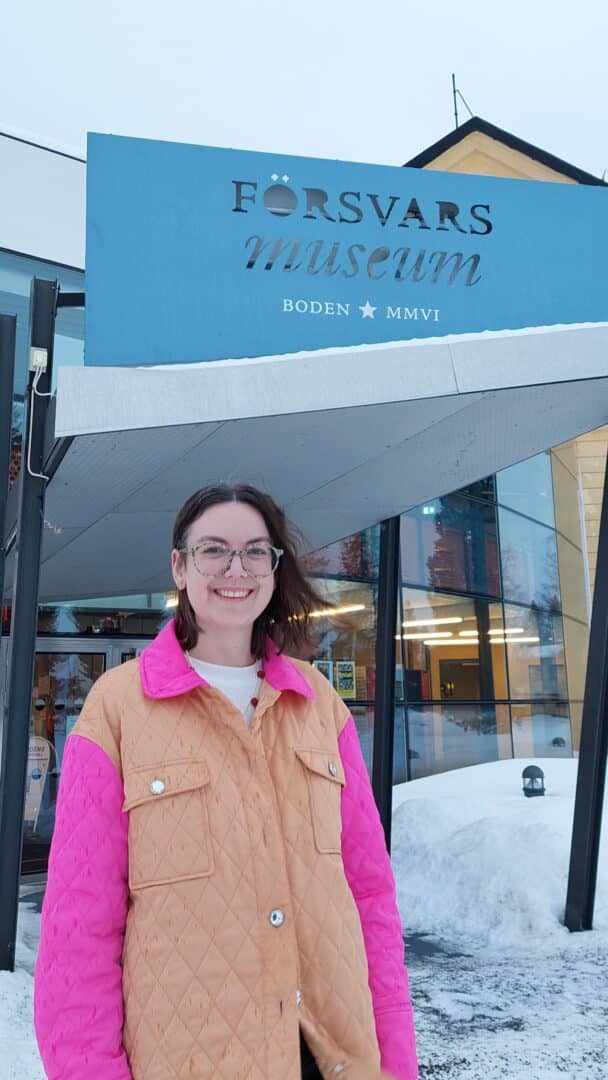Leende kvinna med glasögon och brunt hår, klädd i en rosa och orange quiltad jacka över vit topp, står utomhus framför en glasfasad med snö i bakgrunden.