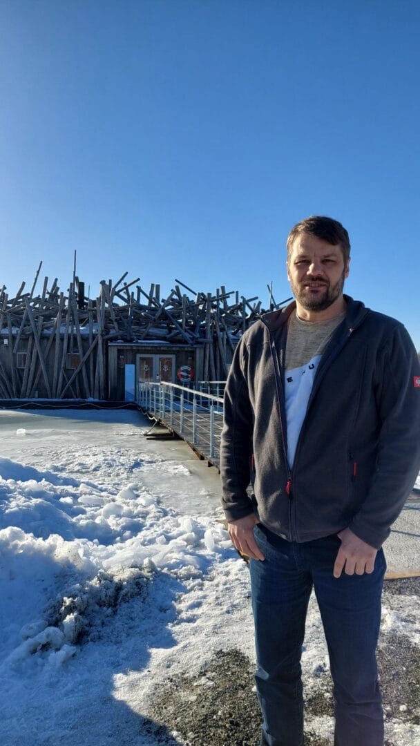 Skäggig man i grå fleecejacka och ljus t-shirt står på en isbelagd brygga framför en träbyggnad med karaktäristisk konstruktion av lutande stockar under klarblå himmel.