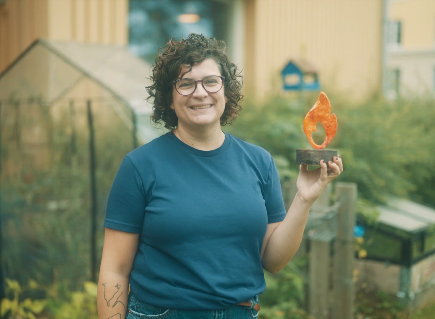 Leende kvinna med mörkt lockigt hår och glasögon i blå t-shirt håller upp en orange flamformad statyett utomhus med växthus och gult hus i bakgrunden.