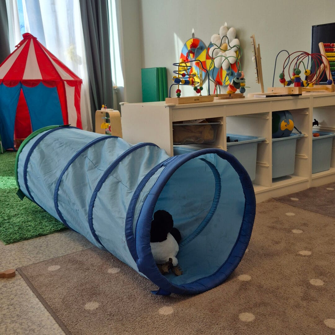 Ett lekrum med leksaker, bland annat en blå tunnel med ett mjukisdjur i, och i bakgrunden olika hjul, pussel och leksakstält.