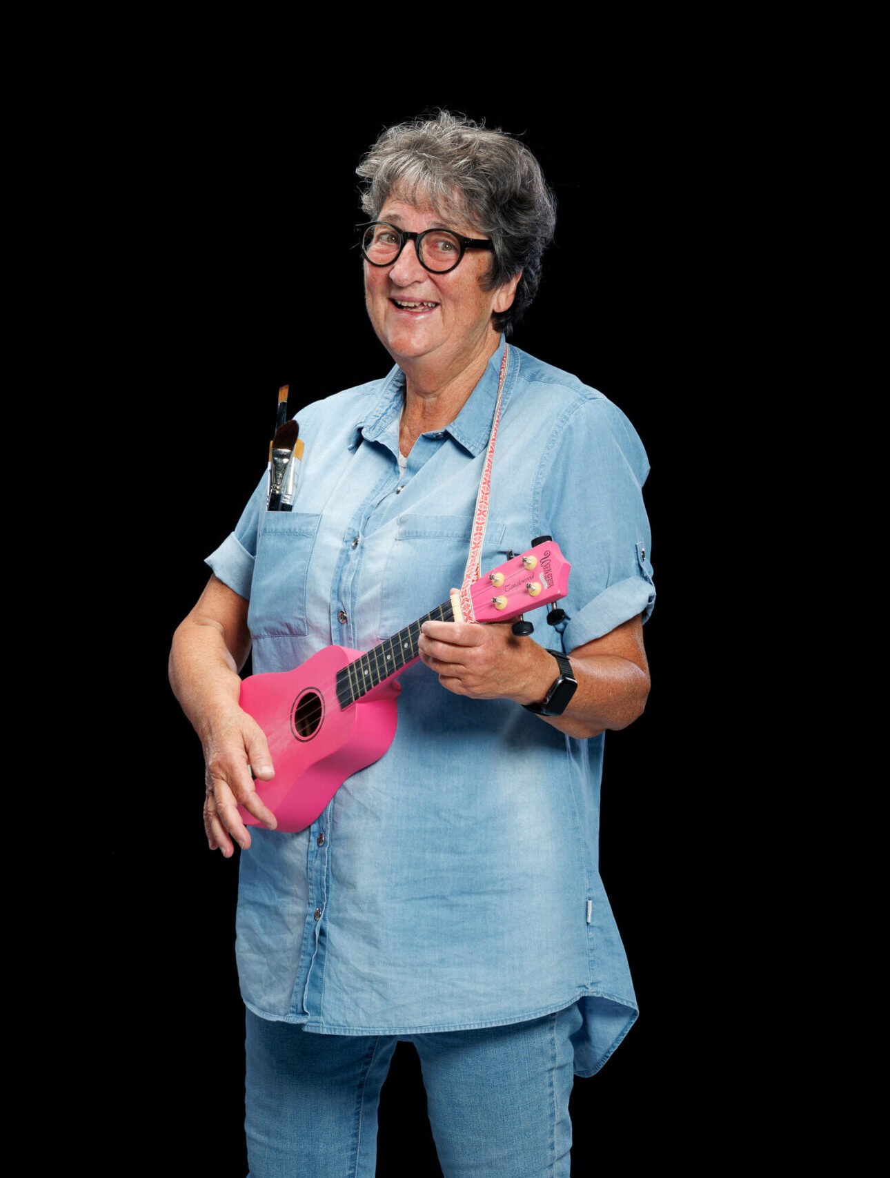 En kvinna står i jeansskjorta framför en svart bakgrund, ler mot kameran och håller i en rosa ukulele