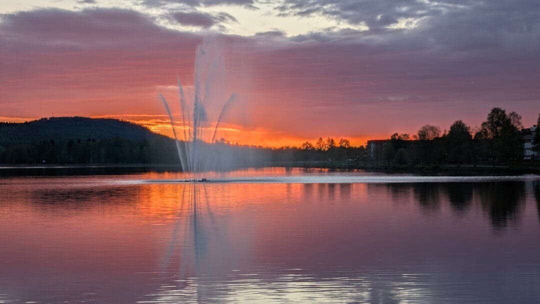 Sunset over a lake with a tall water fountain spraying upward, orange and pink clouds reflecting on the water surface.