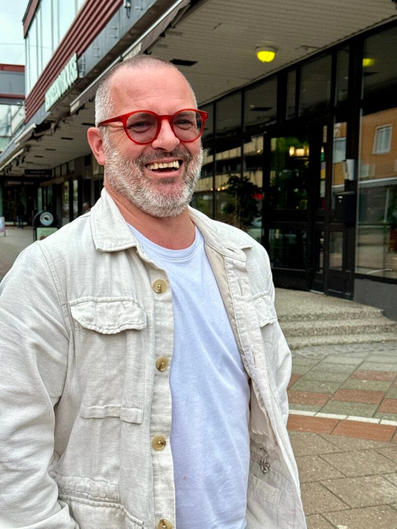 Man with red glasses and a beige jacket smiling at the camera on a city sidewalk outside storefronts.