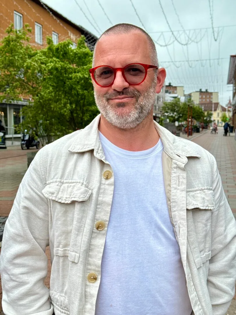 En man med ljus jacka och vit tshirt och röda glasögon står i utomhusmiljö