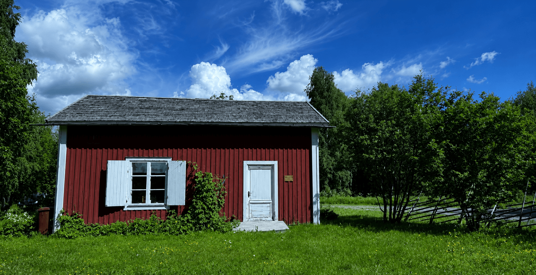 A red wooden cottage with white door and window, set in a grassy yard under a bright blue sky with fluffy clouds.