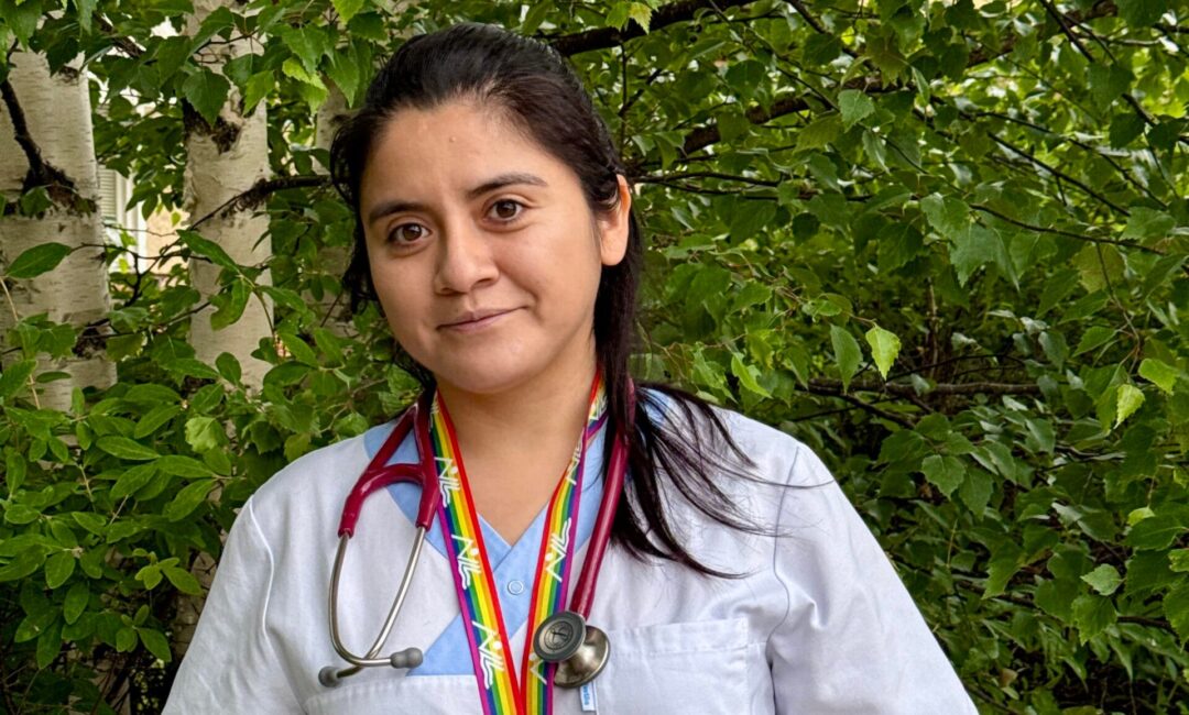 Female healthcare worker in light blue scrubs with stethoscope and ID badge, standing in front of green foliage.